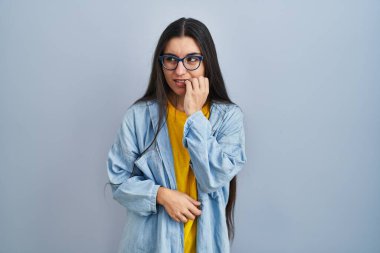 Mavi arka planda dikilen genç İspanyol kadın gergin ve gergin görünüyor. Elleri ağızdan tırnak yiyor. anksiyete. 
