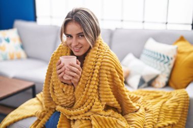 Young blonde woman drinking coffee covering with blanket at home