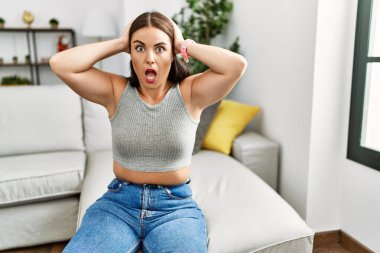 Evdeki kanepede oturan genç esmer kadın çıldırmış ve elleri başının üstünde korkmuş, korkmuş ve ağzı açık şoktan şaşırmış. 