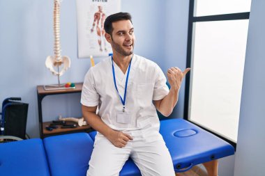 Sakallı, sakallı, acılı bir klinikte çalışan mutlu yüzüyle gülümseyen ve baş parmağıyla yüzünü gösteren genç bir İspanyol.. 