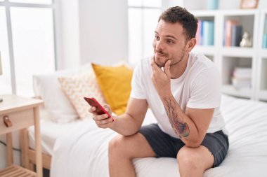 Genç İspanyol adam yatakta akıllı telefon kullanıyor.