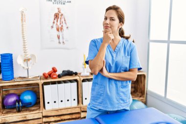 Middle age hispanic physiotherapist woman working at pain recovery clinic looking confident at the camera smiling with crossed arms and hand raised on chin. thinking positive. 