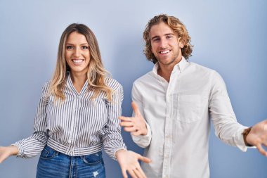 Young couple standing over blue background smiling cheerful with open arms as friendly welcome, positive and confident greetings 