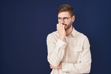 Mavi arka planda duran İspanyol sakallı adam gergin ve gergin görünüyor. Ağzı tırnak ısıran ellerle. anksiyete. 