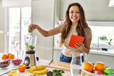Genç, güzel, İspanyol bir kadın blender ile smoothie hazırlıyor. Mutfaktaki tabletten tarifine bakıyor.