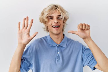 Genç modern adam izole edilmiş arka planda duruyor ve altı numaralı parmaklarıyla işaret ederken kendinden emin ve mutlu bir şekilde gülümsüyor.. 