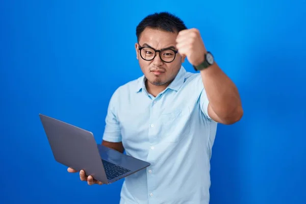 Chinese young man using computer laptop angry and mad raising fist frustrated and furious while shouting with anger. rage and aggressive concept.