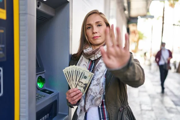 年轻的金发女子拿着自动取款机里的钞票 张开双手 严肃而自信地做着止步手势 摆出一副守口如瓶的架势 — 图库照片