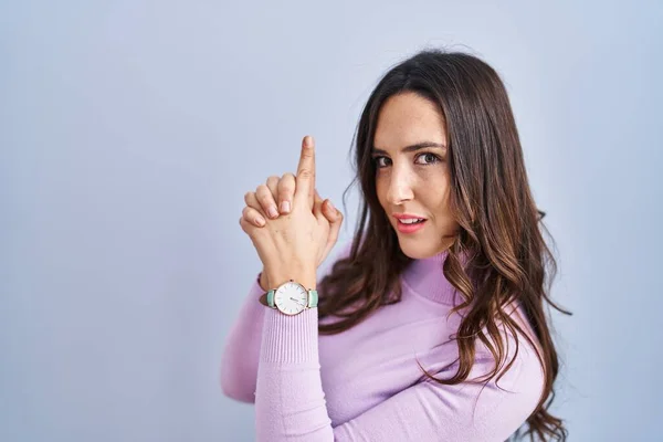 Young brunette woman standing over blue background holding symbolic gun with hand gesture, playing killing shooting weapons, angry face