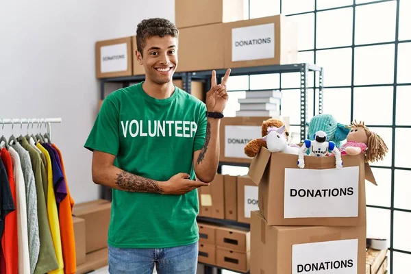 Young handsome hispanic man wearing volunteer t shirt at donations stand smiling with happy face winking at the camera doing victory sign with fingers. number two.