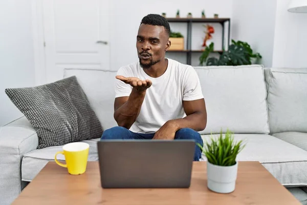 Young african man using laptop at home looking at the camera blowing a kiss with hand on air being lovely and sexy. love expression.