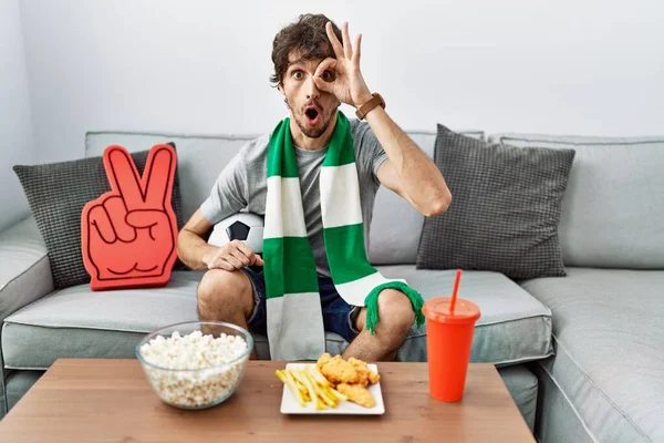 Young hispanic man football hooligan cheering game at home doing ok gesture shocked with surprised face, eye looking through fingers. unbelieving expression.