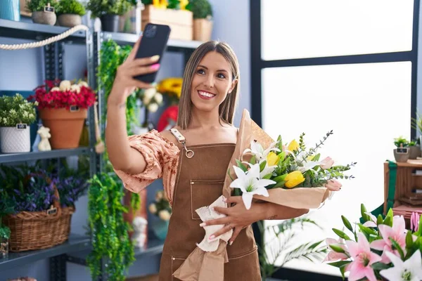 Young beautiful hispanic woman florist make selfie by smartphone holding flowers at flower shop