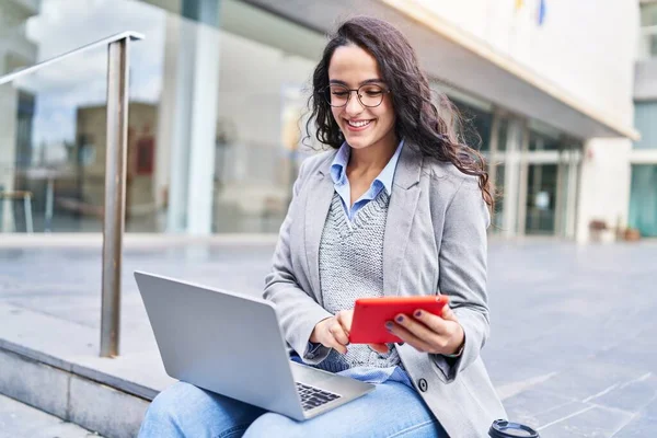 Young hispanic woman executive using laptop and touchpad sitting on bench at street
