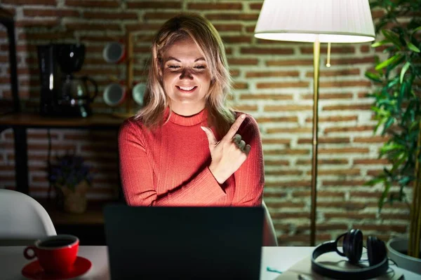 夜に自宅でノートパソコンを使用してブロンドの女性陽気な笑顔で顔を指差す手と指で側に幸せと自然な表情で顔 — ストック写真