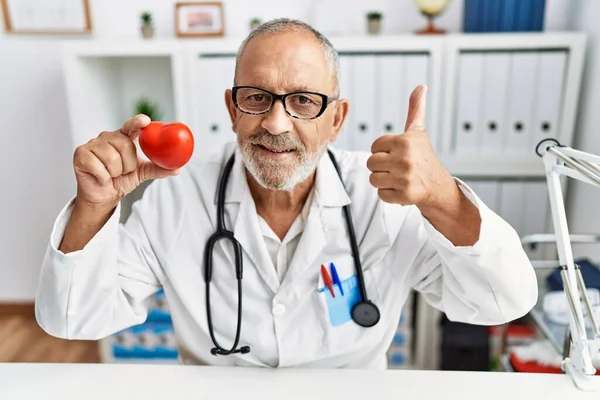 Dojrzały Lekarz Mężczyzna Trzyma Czerwone Serce Klinice Uśmiechając Się Szczęśliwy — Zdjęcie stockowe