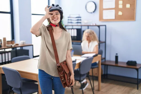 Young brunette woman working at the office wearing bike helmet smiling happy doing ok sign with hand on eye looking through fingers