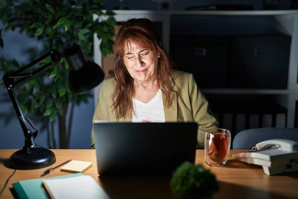 Middle age hispanic woman working using computer laptop at night with hand on stomach because indigestion, painful illness feeling unwell. ache concept.