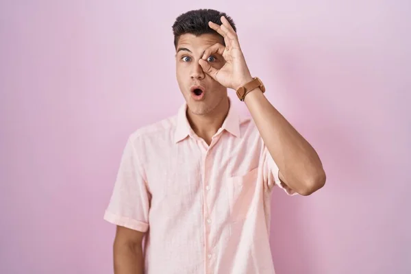 ピンクの背景に立っている若いヒスパニック系の男は驚きの顔でショックを受けたOkジェスチャーを行うと 目の指を見て 信じられない表現 — ストック写真