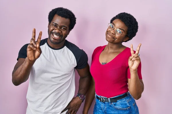 年轻的非洲裔美国夫妇站在粉红的背景上 微笑着看着摄像机 手指头在做胜利的标志 第二点 — 图库照片