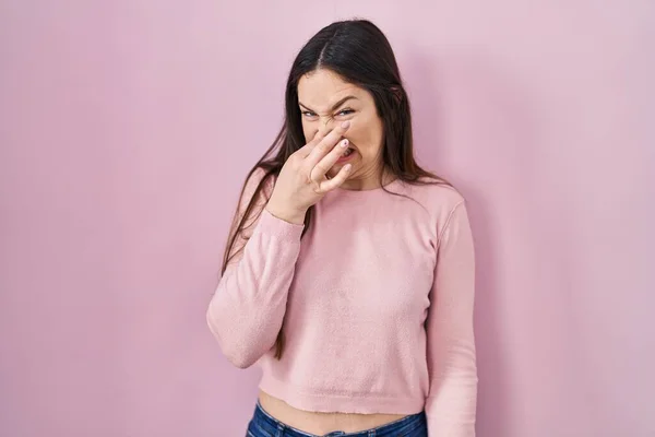 若いブルネットの女性はピンクの背景の上に立っている何か臭いと嫌な 耐えられない匂いを嗅ぎ 鼻の上に指で息を保持する 臭いが悪い — ストック写真