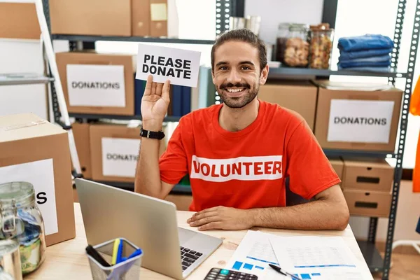 Young hispanic man wearing volunteer t shirt holding please donate banner looking positive and happy standing and smiling with a confident smile showing teeth
