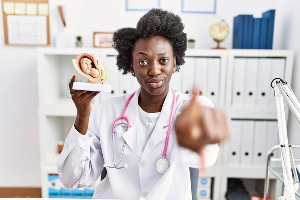African doctor woman holding anatomical model of female uterus with fetus pointing with finger to the camera and to you, confident gesture looking serious