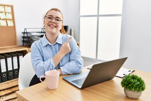 Ofiste çalışan genç, kızıl saçlı bir kadın bilgisayarda neşeli bir dizüstü bilgisayar kullanıyor. Yüzünde, mutlu ve doğal ifadeyle el ve parmak uçlarını gösteren bir gülümseme var. 