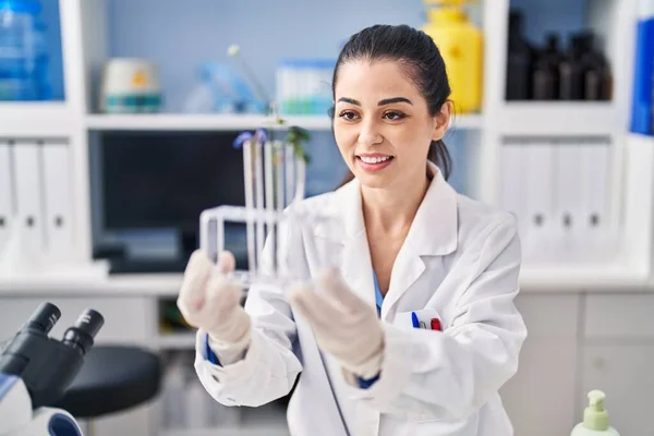 Young beautiful hispanic woman scientist holding test tubes with flowers at laboratory