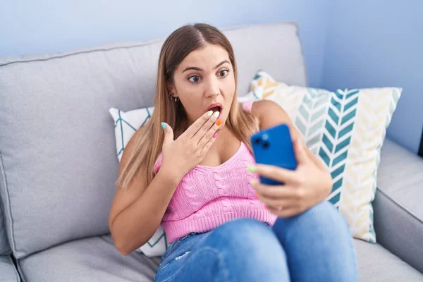 若いブロンドの女性が携帯電話で口を覆い 衝撃を与え 間違いを恐れてソファに座っているスマートフォンでビデオ通話をしています 驚きの表情 — ストック写真