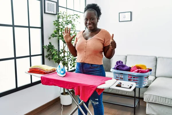 Young african woman ironing clothes at home showing and pointing up with fingers number six while smiling confident and happy.