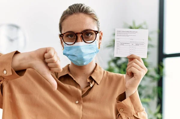 Young blonde woman holding covid record card with angry face, negative sign showing dislike with thumbs down, rejection concept