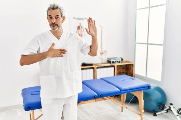 Middle age hispanic therapist man working at pain recovery clinic swearing with hand on chest and open palm, making a loyalty promise oath