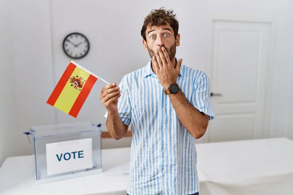 Young handsome man at political campaign election holding spain flag covering mouth with hand, shocked and afraid for mistake. surprised expression