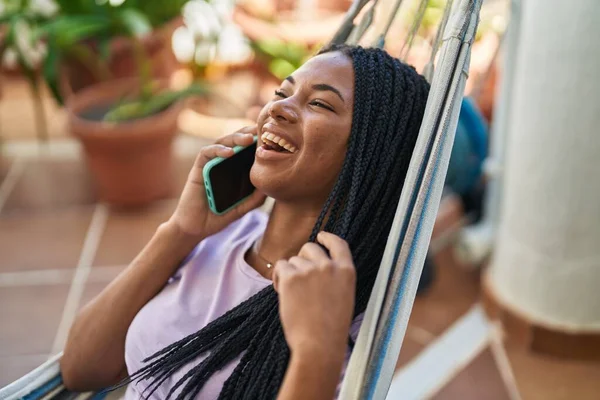 African american woman talking on smartphone lying on hammock at home terrace