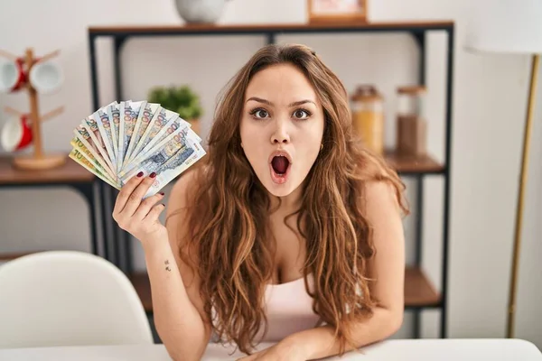 Young hispanic woman holding peruvian sol banknotes scared and amazed with open mouth for surprise, disbelief face