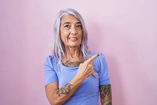 Middle age woman with tattoos standing over pink background pointing aside worried and nervous with forefinger, concerned and surprised expression