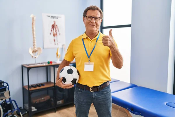 Senior man working at sport physiotherapy clinic smiling happy and positive, thumb up doing excellent and approval sign