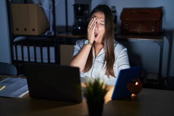 Young brunette woman working at the office at night yawning tired covering half face, eye and mouth with hand. face hurts in pain.