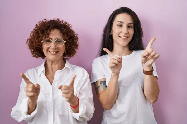 ピンクの背景にカジュアルな白いTシャツを着たヒスパニック系の母親と娘は さまざまな方向に指で自信を持って指して笑っています 広告のコピースペース — ストック写真