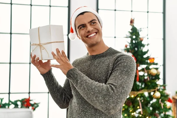 Young Hispanic Man Holding Gift Standing Christmas Tree Home — Φωτογραφία Αρχείου