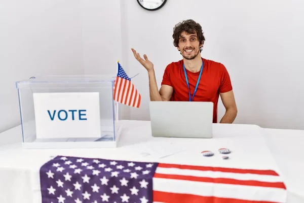 Young hispanic man at political election sitting by ballot smiling cheerful presenting and pointing with palm of hand looking at the camera.