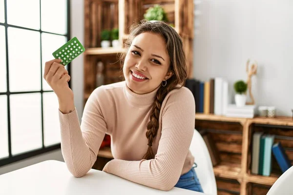 Young beautiful hispanic woman holding birth control pills sitting on table at home