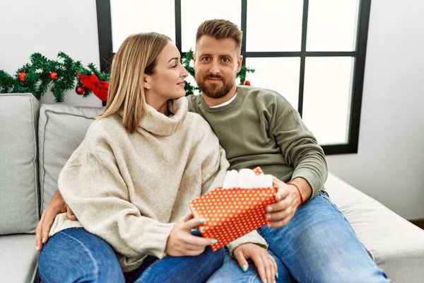 一对年轻的高加索夫妇笑着高高兴兴地抱着礼物坐在家里的圣诞树旁的沙发上 — 图库照片