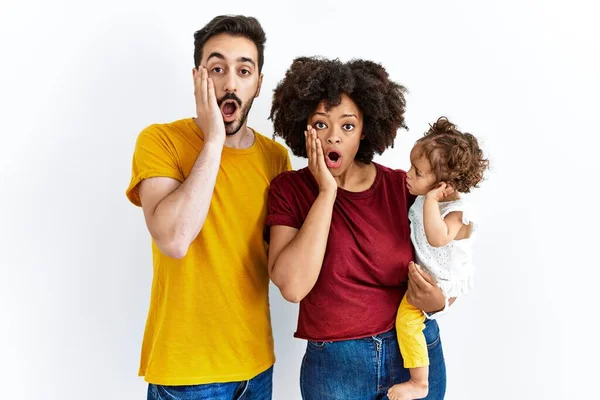 Interracial young family of black mother and hispanic father with daughter afraid and shocked, surprise and amazed expression with hands on face