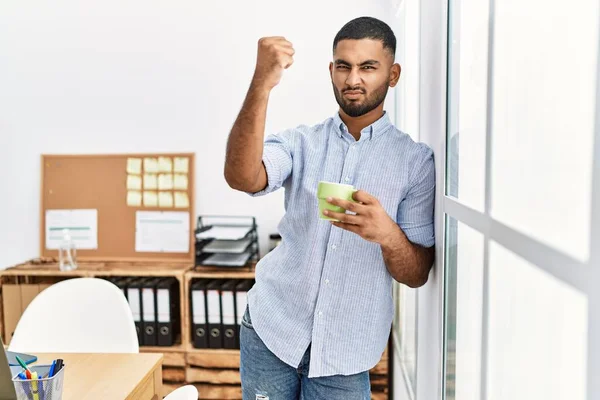 Young indian man drinking a cup coffee at the office annoyed and frustrated shouting with anger, yelling crazy with anger and hand raised