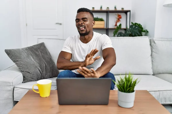Young african man using laptop at home disgusted expression, displeased and fearful doing disgust face because aversion reaction. with hands raised