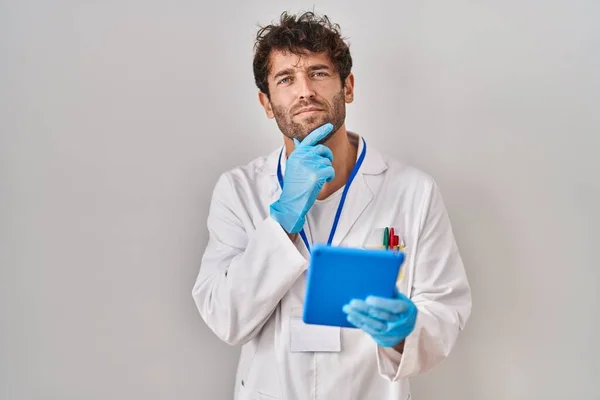 Hispanic scientist man working with tablet serious face thinking about question with hand on chin, thoughtful about confusing idea