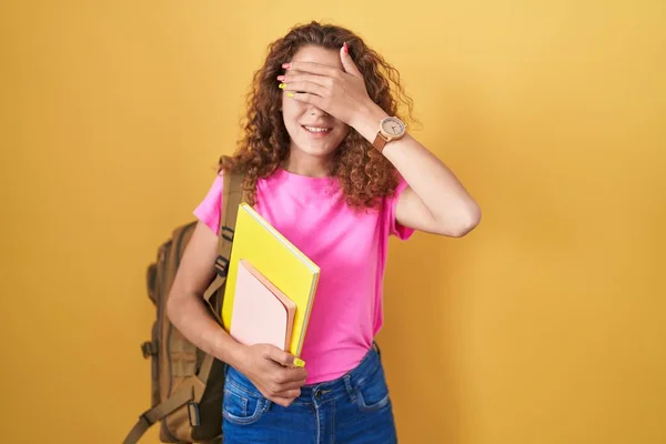 Young caucasian woman wearing student backpack and holding books smiling and laughing with hand on face covering eyes for surprise. blind concept.