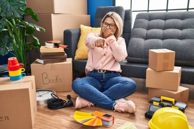 Young hispanic woman moving to a new home sitting on the floor laughing at you, pointing finger to the camera with hand over mouth, shame expression 